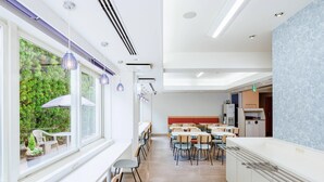 Ένας σύγχρονος χώρος τραπεζαρίας με τραπέζια και καρέκλες, μια κουζίνα με ψυγείο και ένα παράθυρο με θέα σε καταπράσινο περιβάλλον.