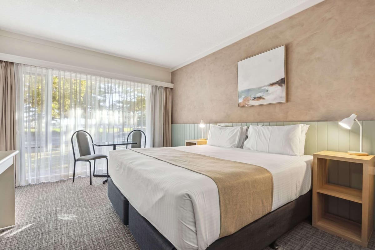 Hotellihuone, jossa on sänky, tuoli, työpöytä, yöpöytä ja taulu seinällä.