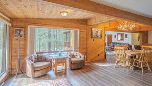 A spacious cabin interior with a wooden floor, a dining area with wooden chairs, a sofa set, and a large window offering a view of the forest. - Byrd Mountain - Upper Canyon Gem - Large Game Room (Ruidoso)