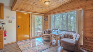 A cozy cabin interior with wooden walls and ceiling, a large window offering a forest view, a fireplace, and a small table with chairs. - Byrd Mountain - Upper Canyon Gem - Large Game Room (Ruidoso)