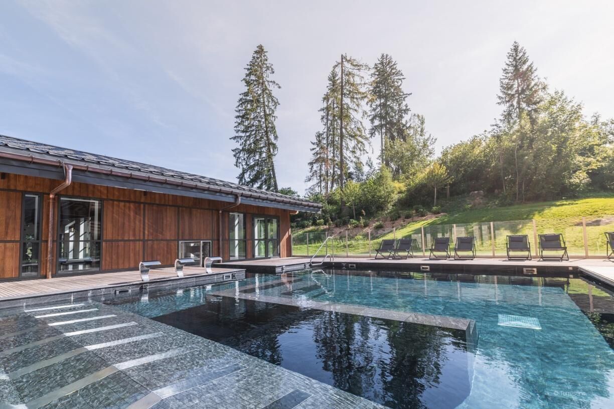 A modern pool area with a wooden deck and lounge chairs, surrounded by tall trees.
