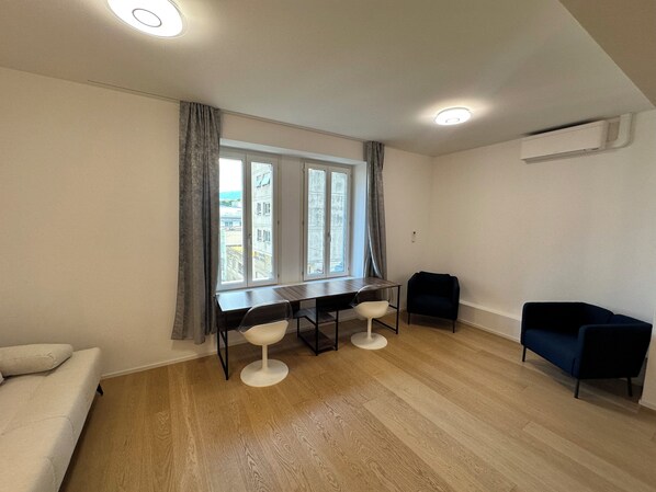 Ein Zimmer mit Holzboden, einem Schreibtisch mit zwei Stühlen, einem Sofa und einem Fenster mit Vorhängen.