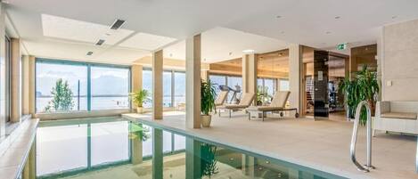 A modern indoor pool area with clear views of the outside, lounge chairs, and a reception desk.