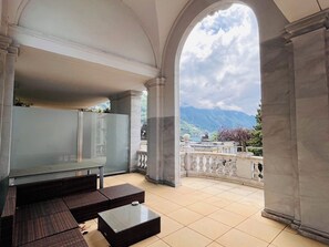 A spacious balcony with a view of mountains, a seating area with wicker furniture, and a marble column structure. - Le National Montreux Switzerland (Montreux)