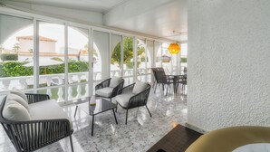 A sunlit patio with a seating area, large windows, and a view of a building in the background.