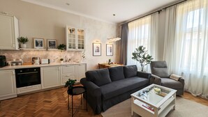 Uma sala de estar moderna com sofá, poltrona e mesa de centro. Há uma área de cozinha com armários e uma mesa de jantar.