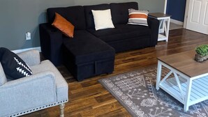 A living room with a black sectional sofa, a grey armchair, and a small white table. There is a framed sign on the wall and a door in the background. - Cozy building in charming Loretto with WiFi, AC (Loretto)