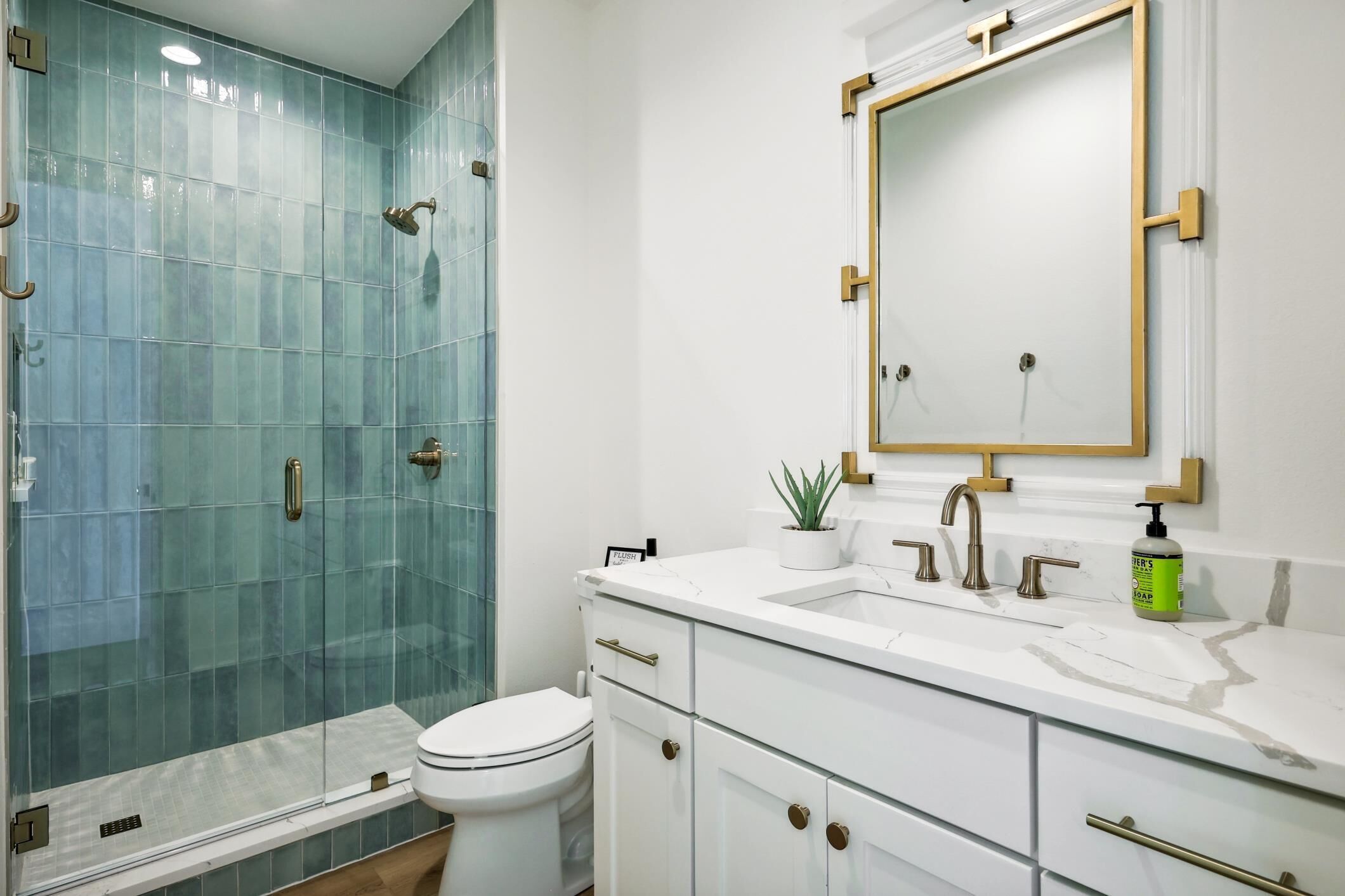 A modern bathroom with a glass shower enclosure, a white toilet, a marble sink with gold fixtures, and a large mirror with a gold frame.