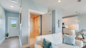 A modern living room with a white sofa, teal cushions, and a view of a kitchen and balcony. - Cape Ocean Front~Jacuzzi~Elevator~Arcade Games! (Port St. Joe)
