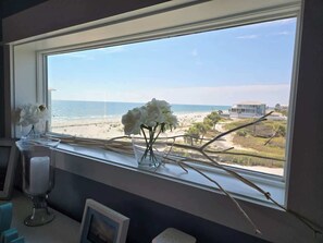 A window with a view of the beach and ocean, a vase with white flowers, and a candle holder.