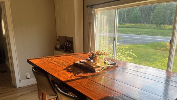 A wooden dining table with chairs, a window with a view of a road and greenery, and a shelf with bottles.