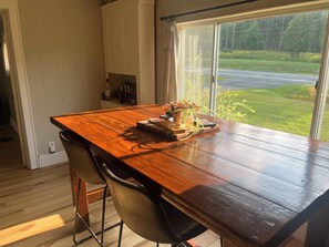 A wooden dining table with chairs, a window with a view of a road and greenery, and a shelf with bottles.