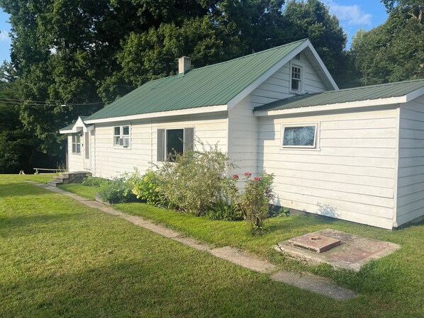 A white wooden house with a green roof, a front porch, and a small garden with pink flowers.