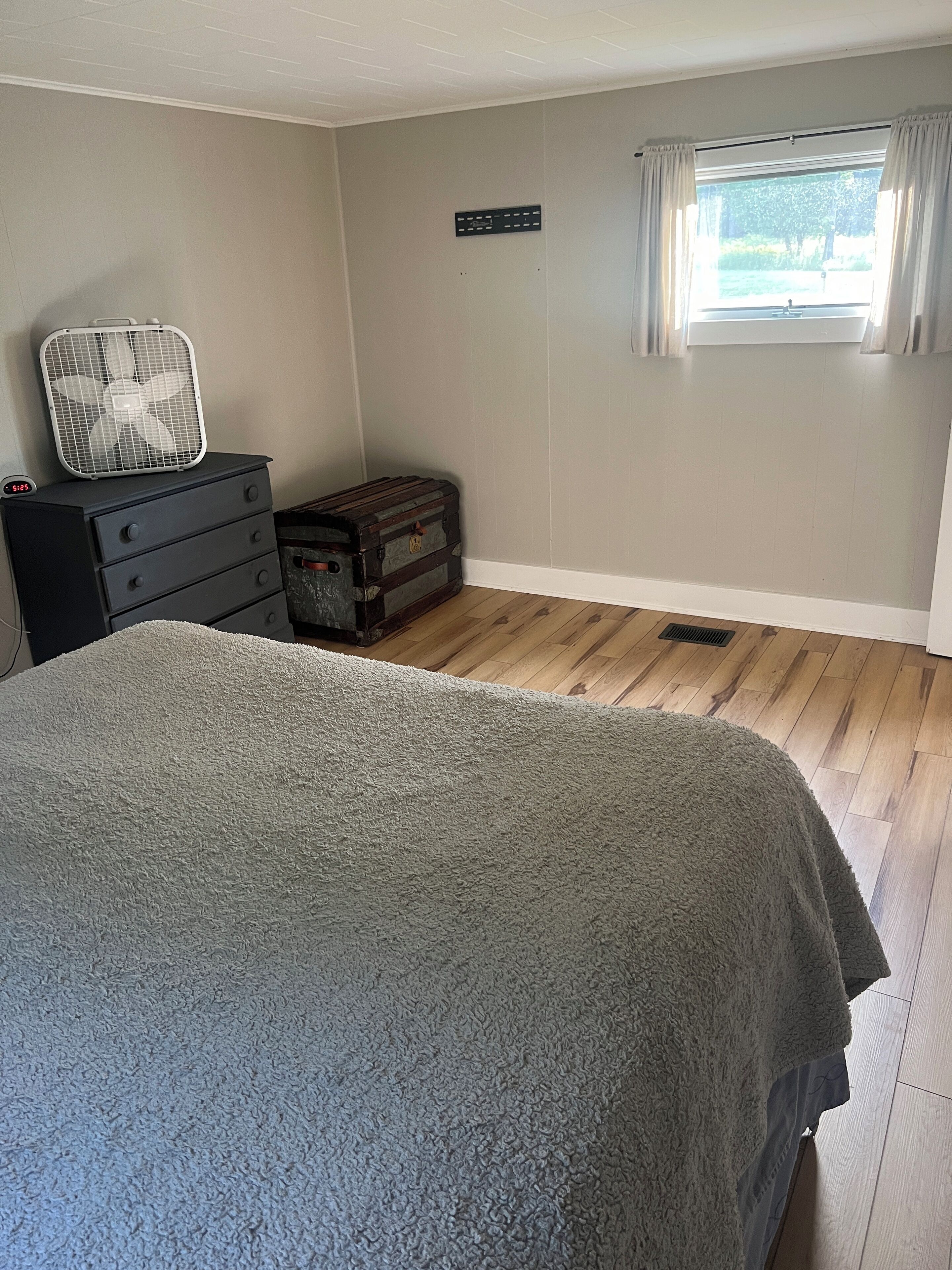 A bedroom with a large bed, a dresser, a suitcase, a fan, and a window with curtains.