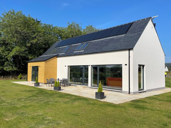 Une maison moderne dotée d’un toit en pente, de grandes fenêtres et d’un patio aménagé avec du mobilier d’extérieur.