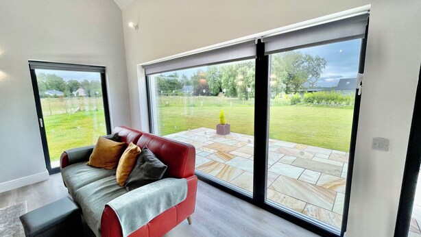 A modern living room with a red sofa, a grey throw, and a view of a garden through large sliding glass doors. - Ash Mill - Dornoch (Dornoch)