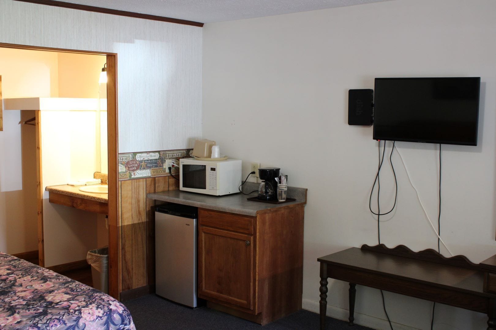 A compact hotel room with a kitchenette, a microwave, a coffee maker, a small refrigerator, a TV, and a bed with a floral bedspread.