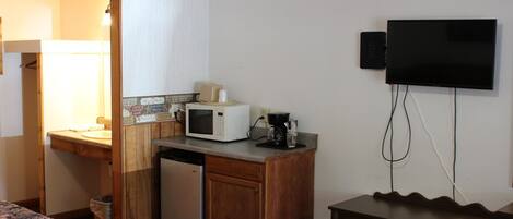 A compact hotel room with a kitchenette, a microwave, a coffee maker, a small refrigerator, a TV, and a bed with a floral bedspread.