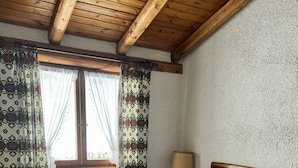 A bedroom with a wooden ceiling, a bed with a white duvet, a nightstand with a lamp, and a window with patterned curtains. - Appartamento Marele (Folgarida)