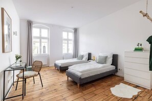 A modern bedroom with two single beds, a wooden floor, a desk with a chair, and a large window.