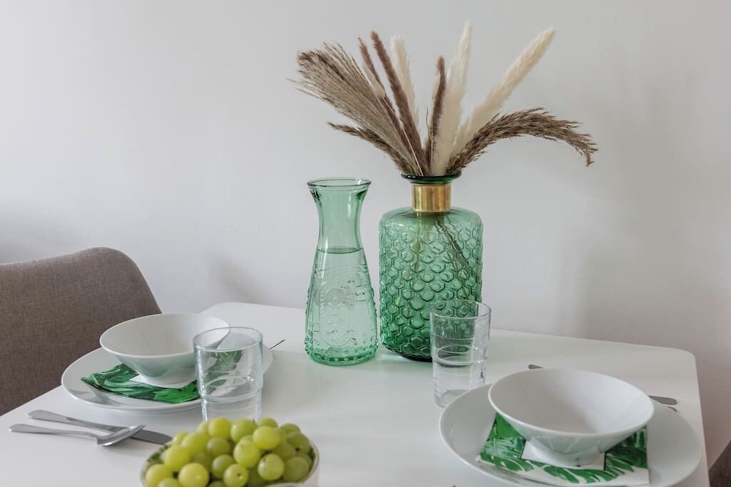A dining table with a white tablecloth, two green glass vases, a bowl of grapes, and a white bowl on a plate.
