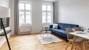 Ein modernes Wohnzimmer mit einem blauen Sofa, einem Esstisch mit Stühlen und einem kleinen Couchtisch. Zwei Fenster mit Vorhängen, ein Heizkörper und eine Lampe sind vorhanden.