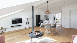Un vaste espace de vie doté d’un haut plafond, d’un téléviseur à écran plat fixé au mur, d’un coin repas avec une table et des chaises, et d’une cuisine située à l’arrière-plan.