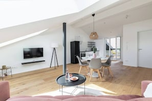 A spacious living area with a high ceiling, a flat-screen TV mounted on the wall, a dining area with a table and chairs, and a kitchen in the background.
