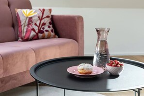 A pink sofa with a colorful pillow, a black coffee table with a donut, a glass pitcher, and a bowl of strawberries.