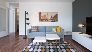 A modern living room with a sofa, two white coffee tables, a patterned rug, and a cityscape painting on the wall. - 65 m² Apartment w. Spreeview directly at Ostkreuz (Berlin)