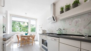 A modern kitchen with white cabinets, a wooden dining table, and a window with a view of greenery. - 65 m² Apartment w. Spreeview directly at Ostkreuz (Berlin)