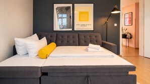 A modern bedroom with a large bed, a dark grey upholstered headboard, and two framed artworks on the wall.