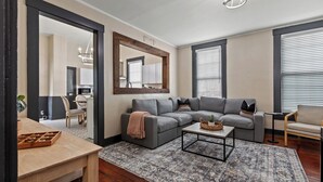 Una sala de estar con un sofá seccional gris, una mesa de centro y un tapete estampado. Se ve una mesa de comedor de madera con sillas, un espejo y un área de cocina a través de una puerta abierta.