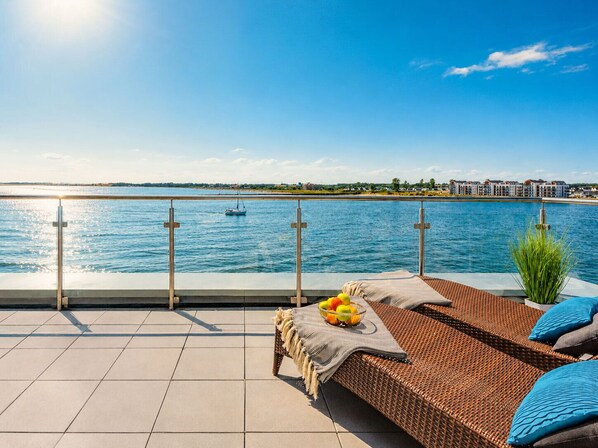 En terrass med utsikt över havet, en loungehörna med en flätad soffa, en skål med frukt och ett räcke.
