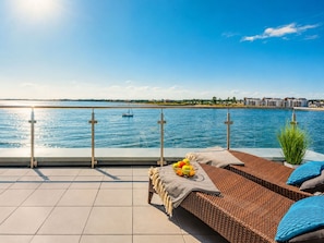 Una terraza con vista al mar, un área de descanso con un sofá de mimbre, un cuenco de frutas y una barandilla.