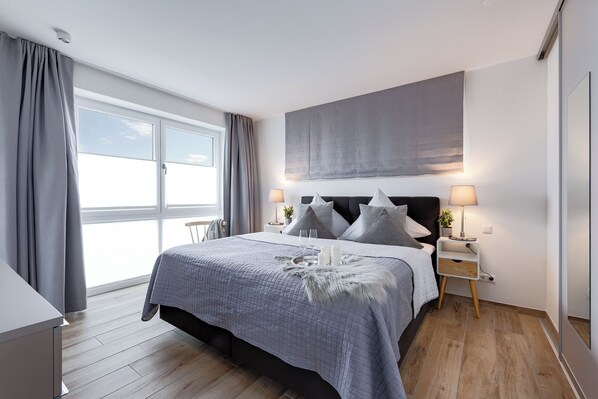 A modern bedroom with a large bed, two bedside tables with lamps, a grey headboard, and a view of the sky through a large window. - "Sky 65" - Exclusive penthouse with sauna & sea view at Ostsee Resort Olpenitz (Ostseeresort Olpenitz)