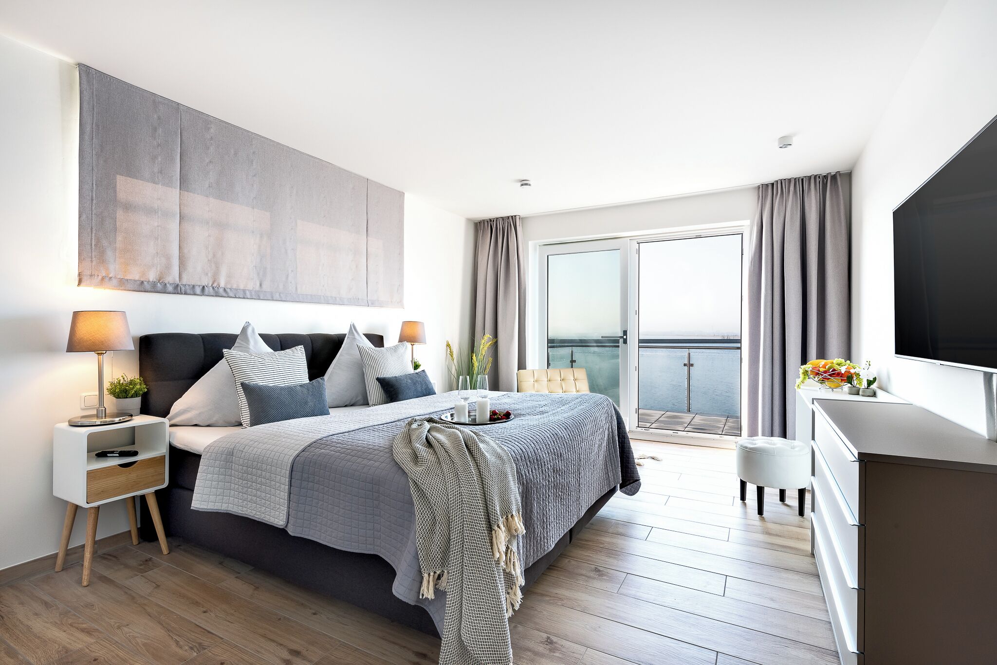 Ein modernes Schlafzimmer mit einem großen Bett, Nachttischen, einem Fernseher und einem Balkon mit Vorhängen.