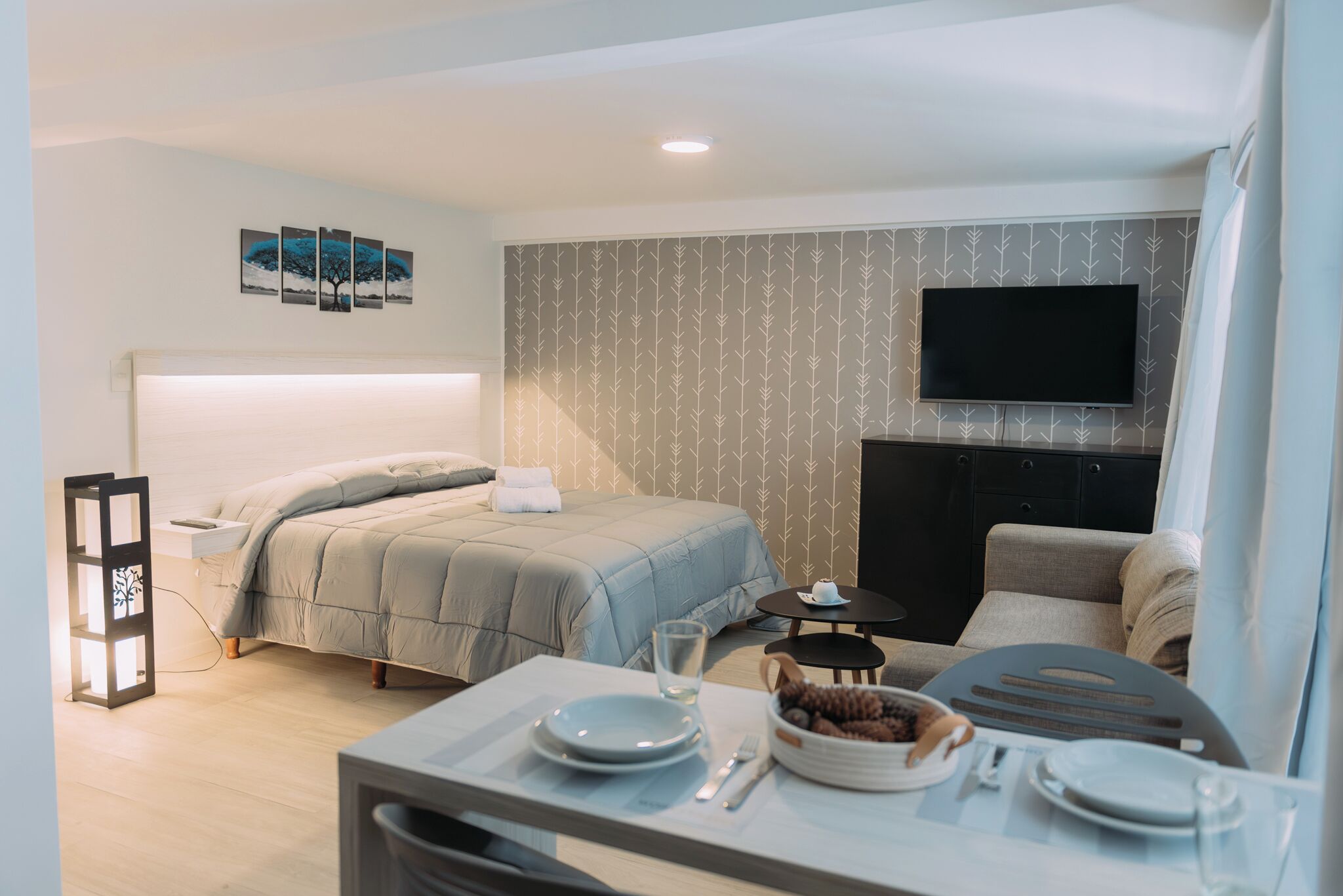 A modern hotel room with a bed, bedside table, TV, and a dining area with a table set for two.