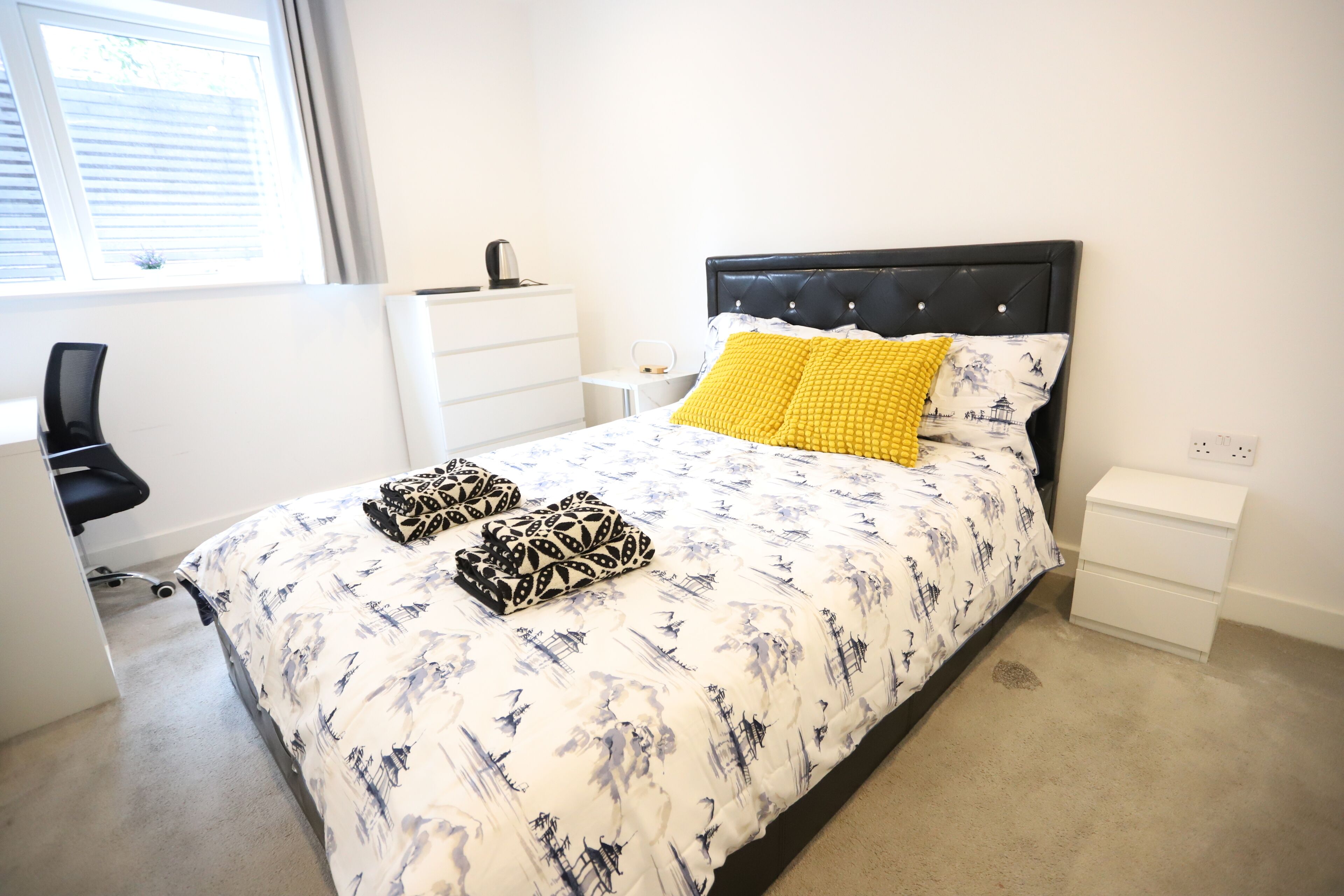A bedroom with a bed, two pillows, a bedside table, a chair, a desk, and a window.
