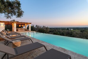 Ein Poolbereich mit Liegestühlen und einer Pergola mit Beleuchtung, mit Blick auf eine Landschaft mit Bäumen und eine ferne Aussicht.