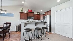 A kitchen with wooden cabinets, a stainless steel refrigerator, a microwave, and a sink. There are three stools and a dining table with chairs.