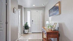 A hallway with a white door, a welcome sign, a potted plant, and a wooden table with a lamp and picture frame.