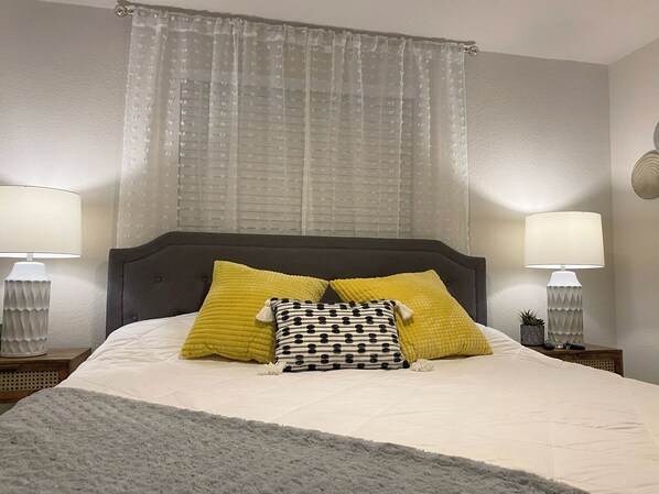 Una cama bien tendida con un edredón gris, dos lámparas de noche y una ventana con cortinas traslúcidas.