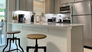 A modern kitchen with grey cabinets, a stainless steel refrigerator, a microwave, and a coffee maker. There is a small island with stools and a potted plant.