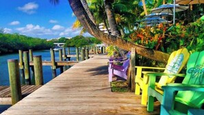 A wooden pier extending into a body of water, with colorful chairs and a hammock under a palm tree.