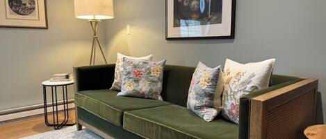 Un sofá verde con cojines florales, una lámpara de pie, una obra de arte floral enmarco y una mesita con libros.