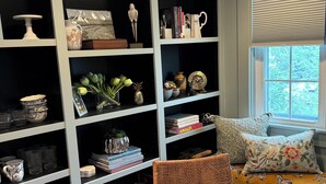 A room with a large bookshelf filled with books, decorative items, and a dining area with a round table and wicker chairs.