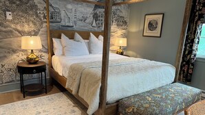 A four-poster canopy bed with a white bedspread and pillows, flanked by two bedside tables with lamps, a patterned rug, and a bench with a floral upholstery.