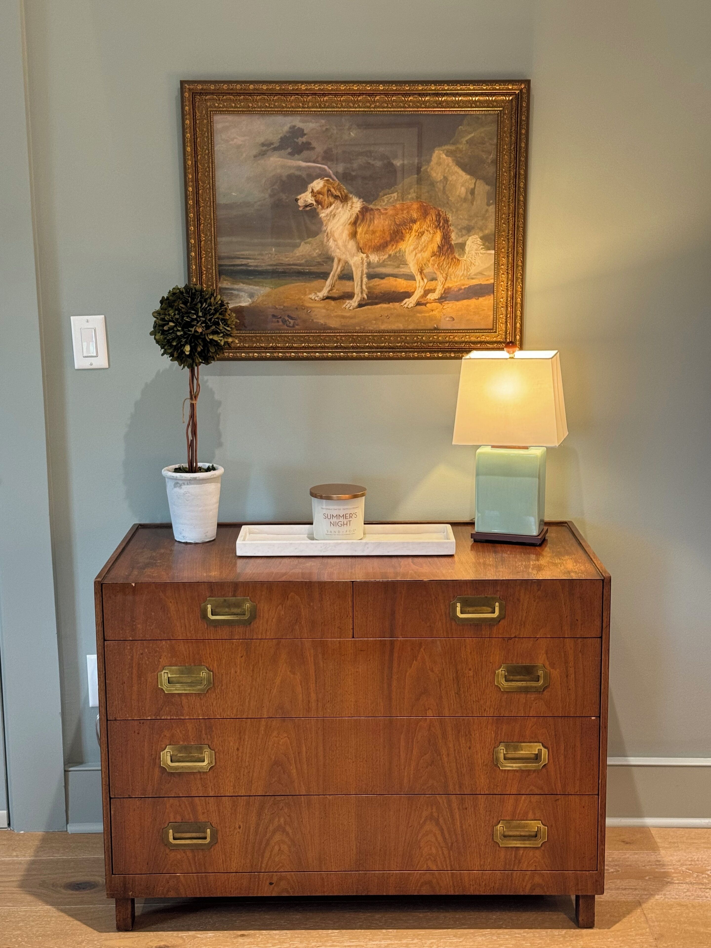 A wooden dresser with brass handles, a framed painting of a dog, a potted plant, and a candle on top.