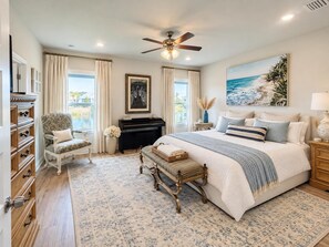 A bedroom with a large bed, a ceiling fan, a piano, a chair, and a view of the outdoors.